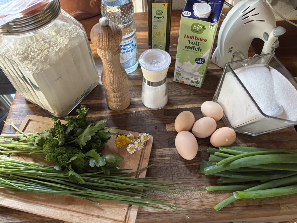 Zutaten für die Pfannkuchen bereitgestellt, Mehl, Kohlensäurehaltiges Wasser, neutrales Öl zum ausbacken, Milch, Handrührgerät, Eier, Zucker, Salz und Pfeffer und Kräuter wie Winterheckenzwiebel, Petersilie, Schnittlauch, Gänseblümchen und Löwenzahn